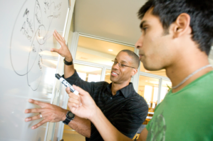 A person is explaining a concept on a whiteboard to another person, holding a marker. This scene represents a collaborative learning or teaching environment.