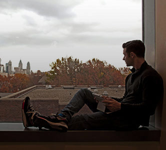 A person sits on a windowsill, holding a book and gazing out at a city skyline with autumn trees in the foreground, under a cloudy sky.